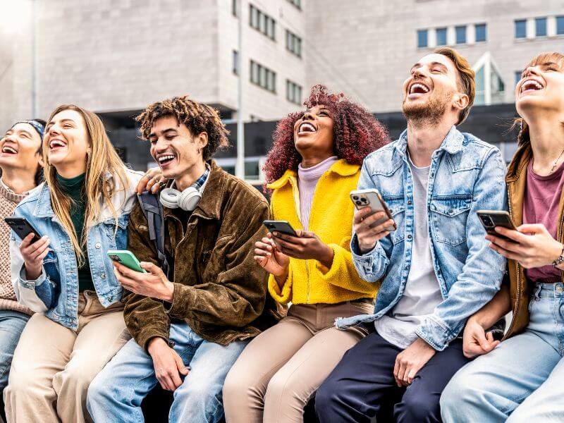 Group of young adults laughing together outdoors with their phones